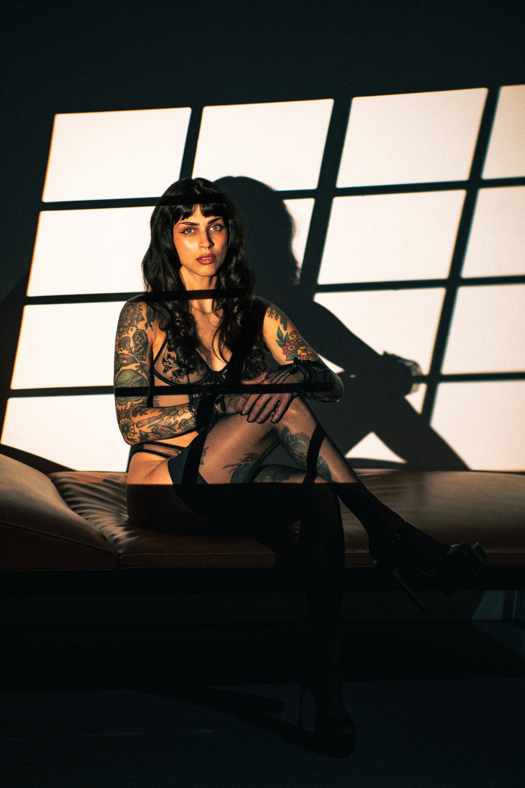 A tattooed woman sits on a leather lounge chair in a dimly lit room, wearing sheer black stockings and lingerie, with dramatic window light casting strong shadows behind her.
