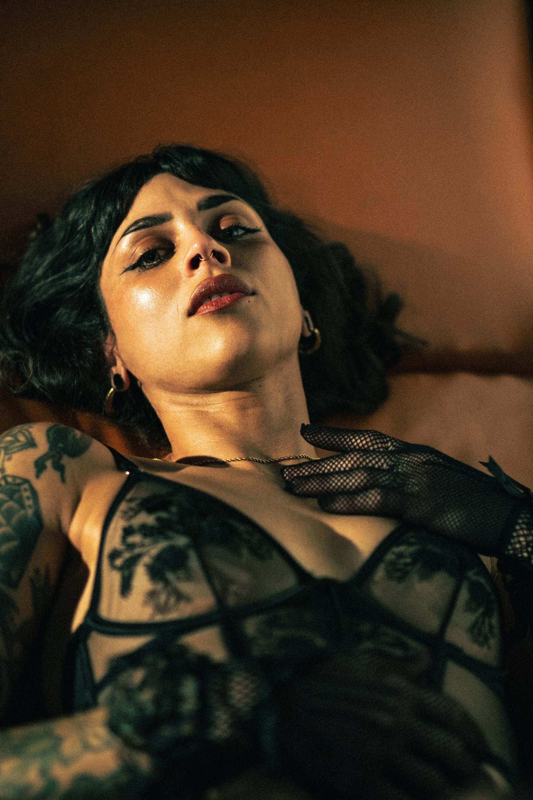 A woman with short dark hair and visible tattoos reclines against a warm brown background, wearing a sheer black lace lingerie top and black fishnet gloves.