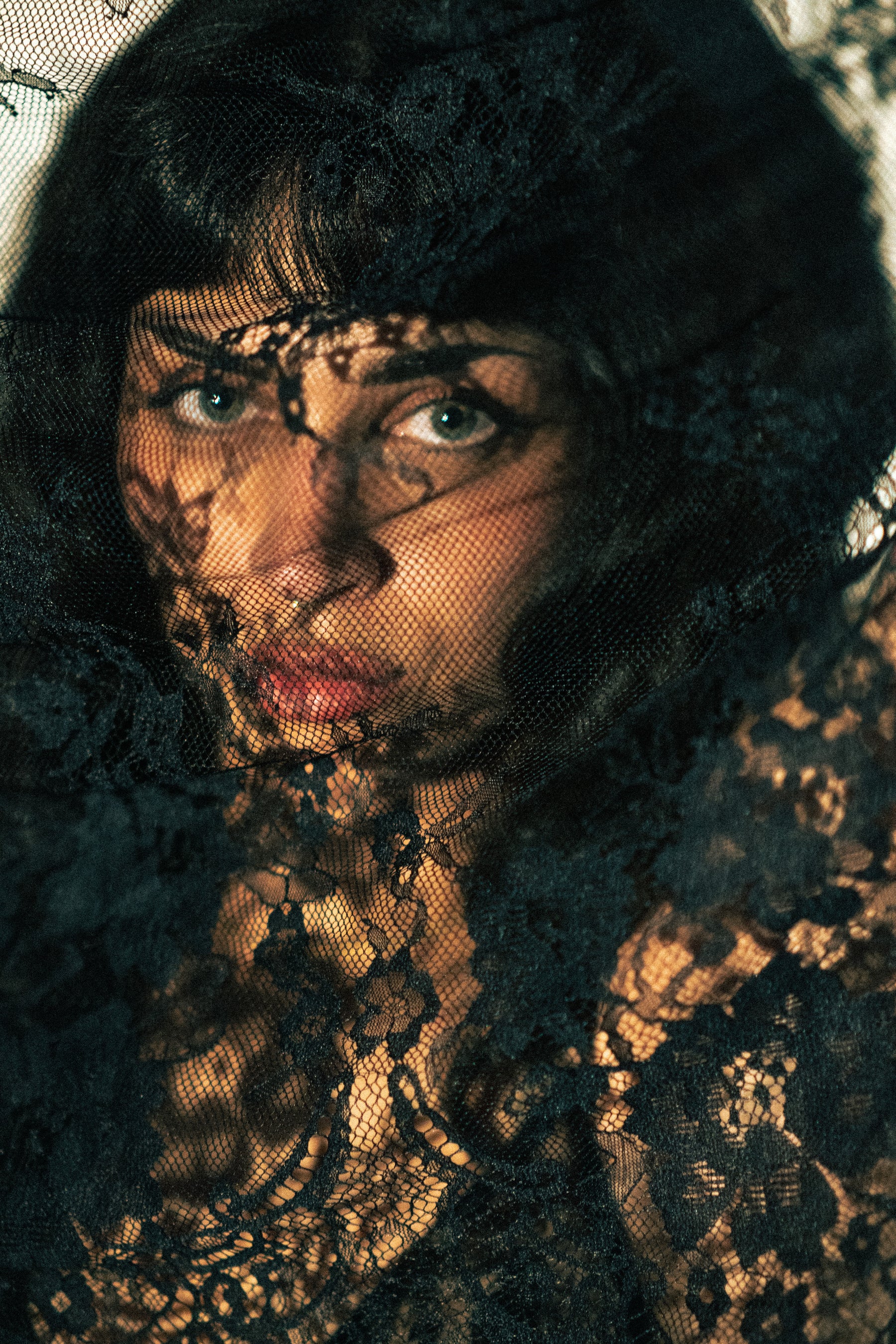A close-up portrait of a woman wearing a sheer black lace veil over her face and a black lace garment, with warm golden light highlighting the lace texture.