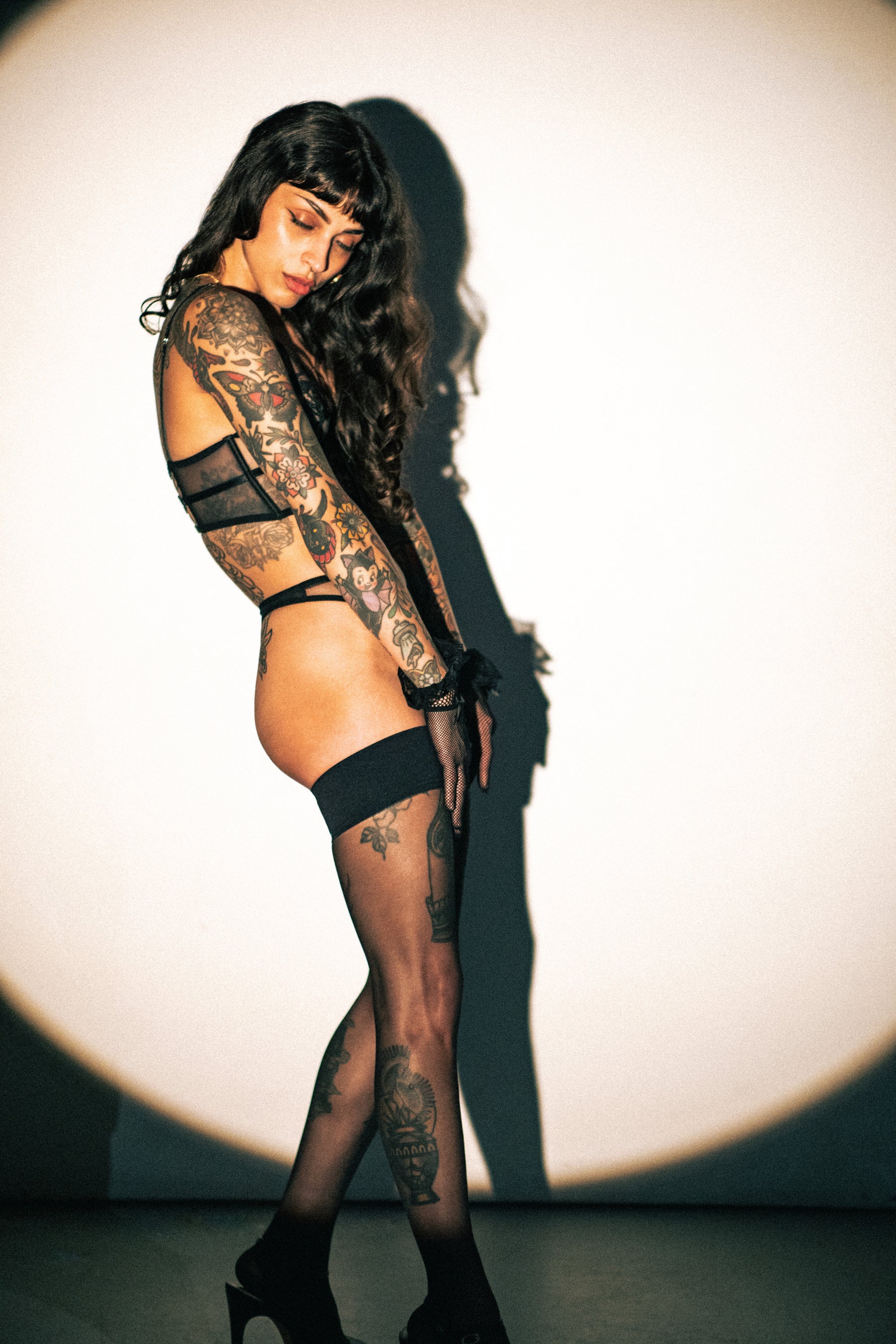 A tattooed woman poses in profile wearing sheer black thigh-high stockings, a black strappy lace bodysuit, and black high heels against a white studio background with a dramatic spotlight and shadow.