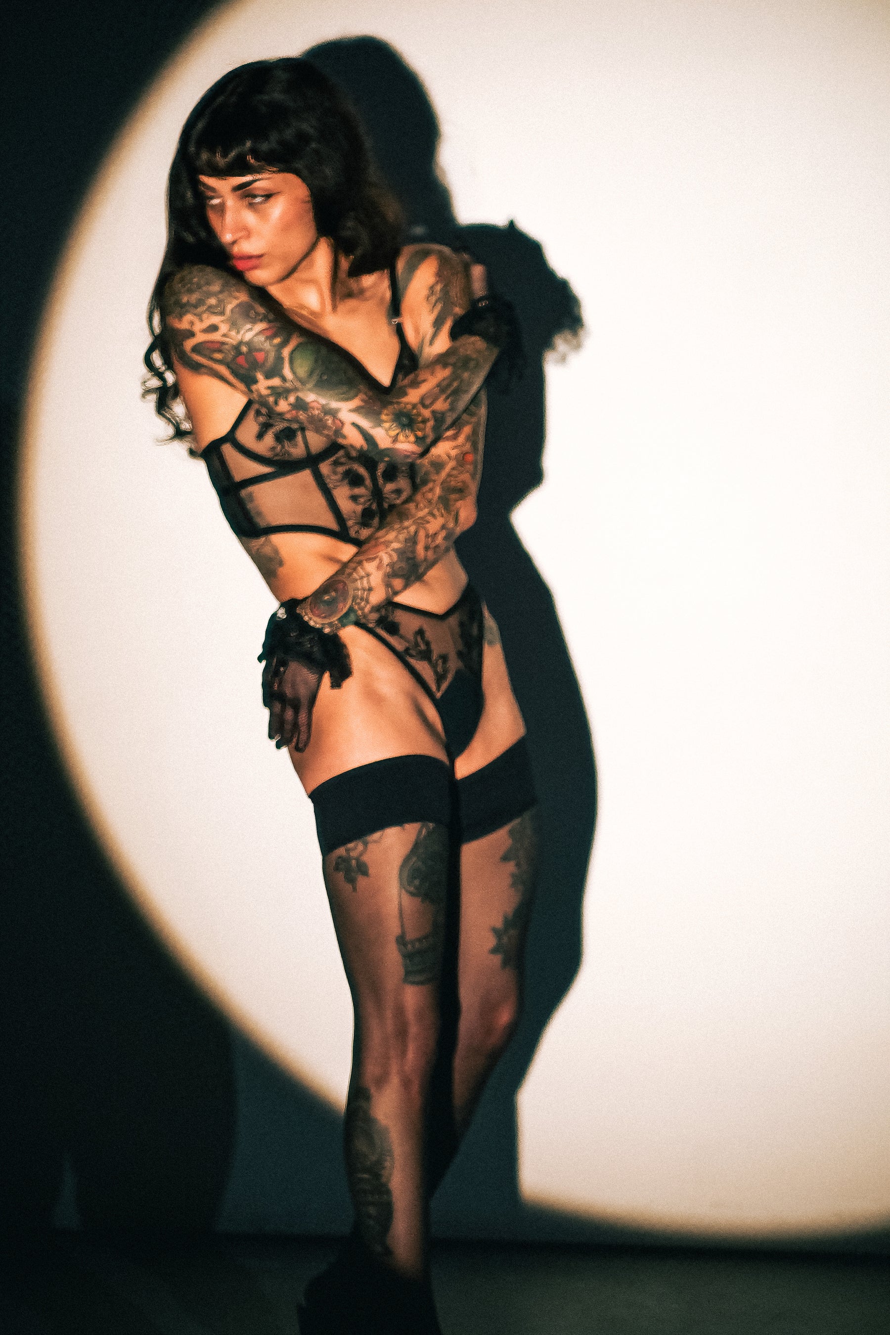 A tattooed woman poses in a black strappy lingerie bodysuit with matching thigh-high stockings against a spotlighted white background.