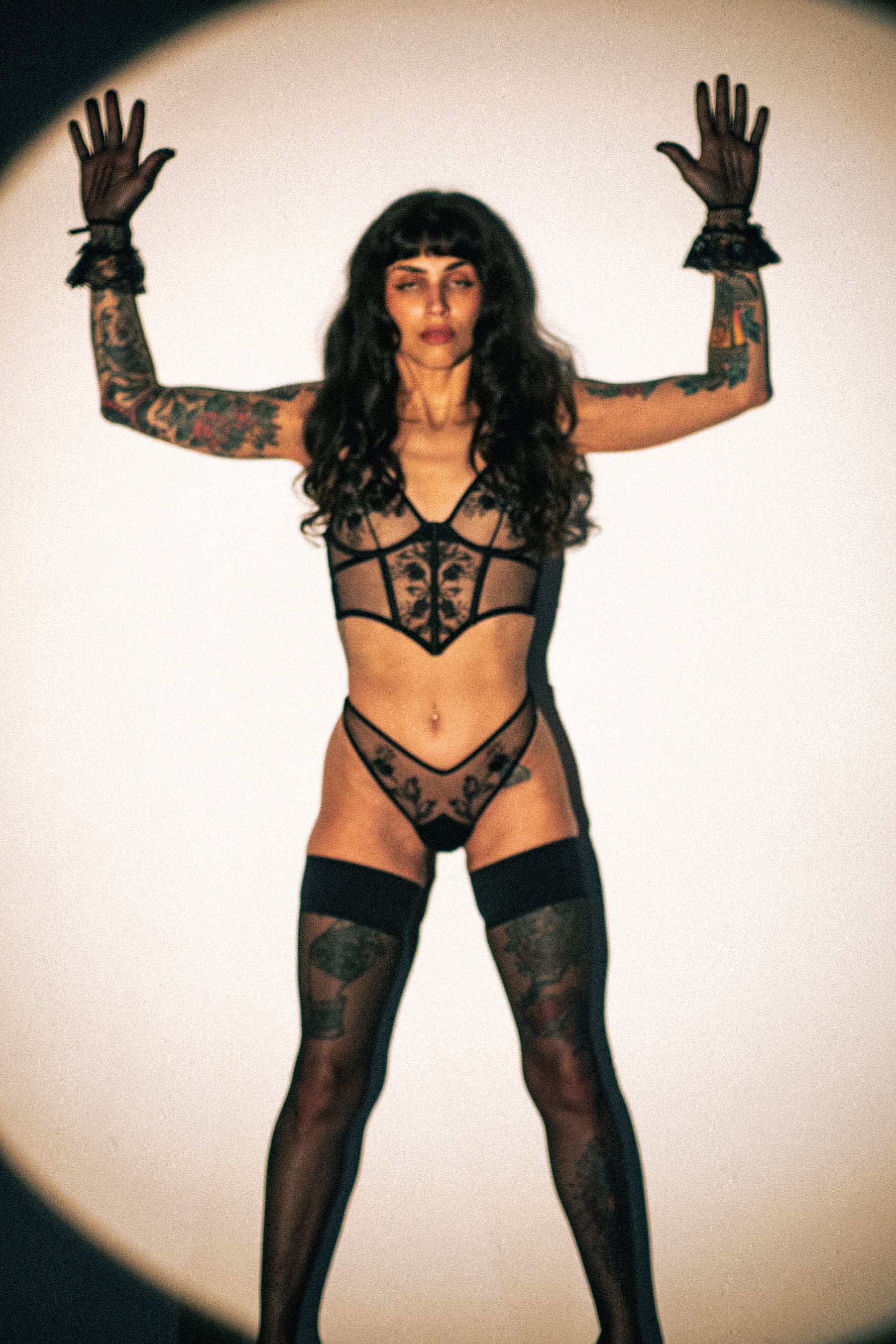 A model with long dark hair and visible tattoos poses with arms raised, wearing a black strappy lingerie set, sheer black thigh-high stockings, and black gloves against a circular spotlight on a white background.