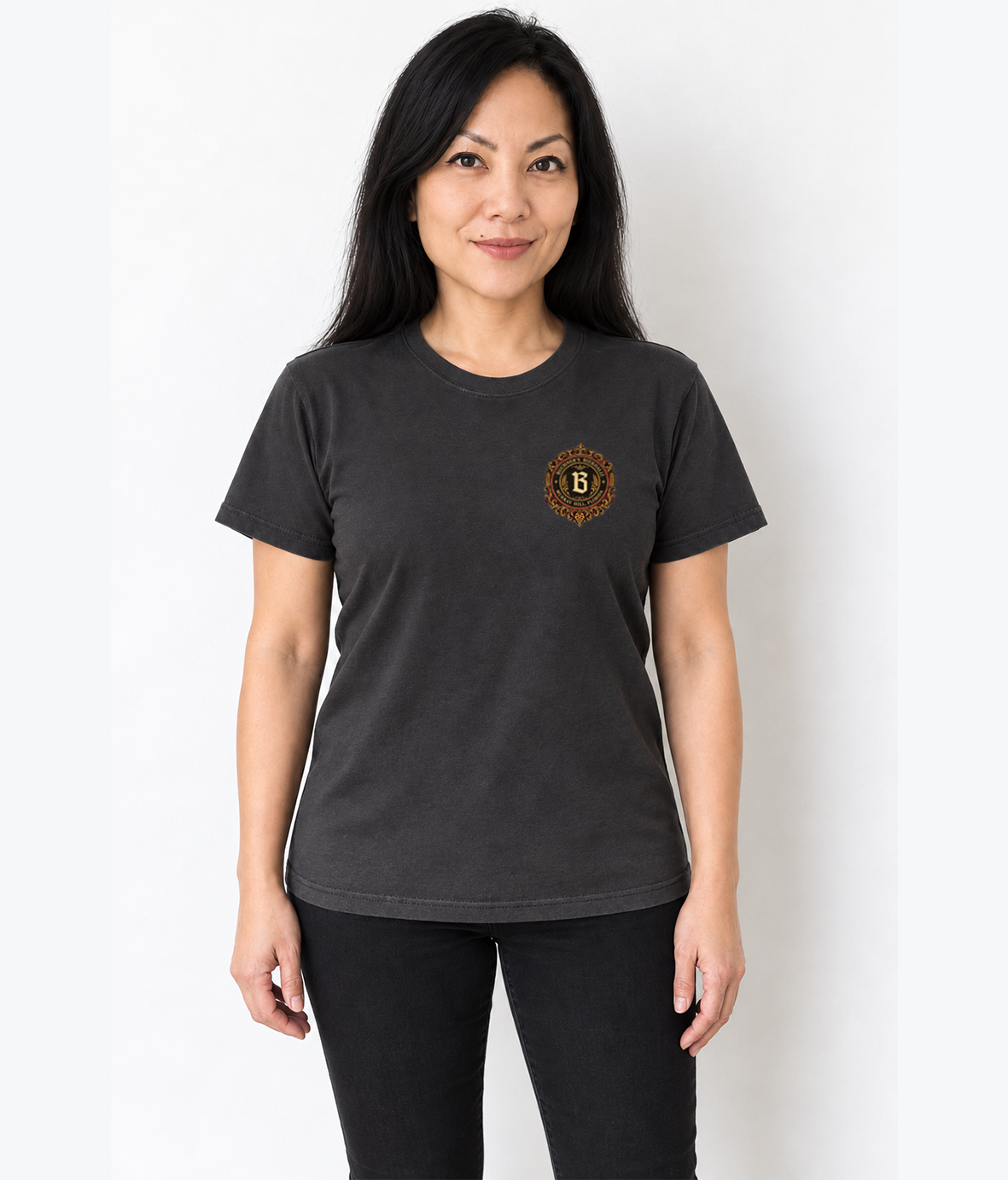 A woman modeling a dark charcoal gray short-sleeve t-shirt with a small circular gold and black graphic on the left chest, paired with black pants, against a plain white background.