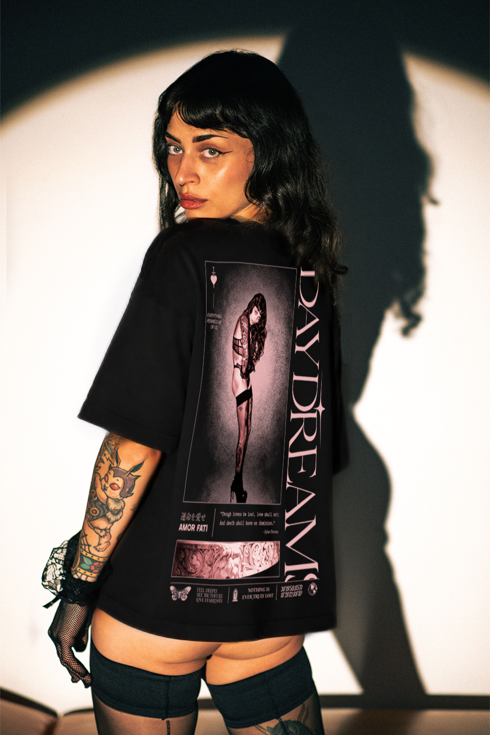 A model wearing a black graphic t-shirt with a large back print featuring a gothic woman and the text 'DAYDREAM' and 'MORNING NIT', shown from behind against a dramatic spotlight background.
