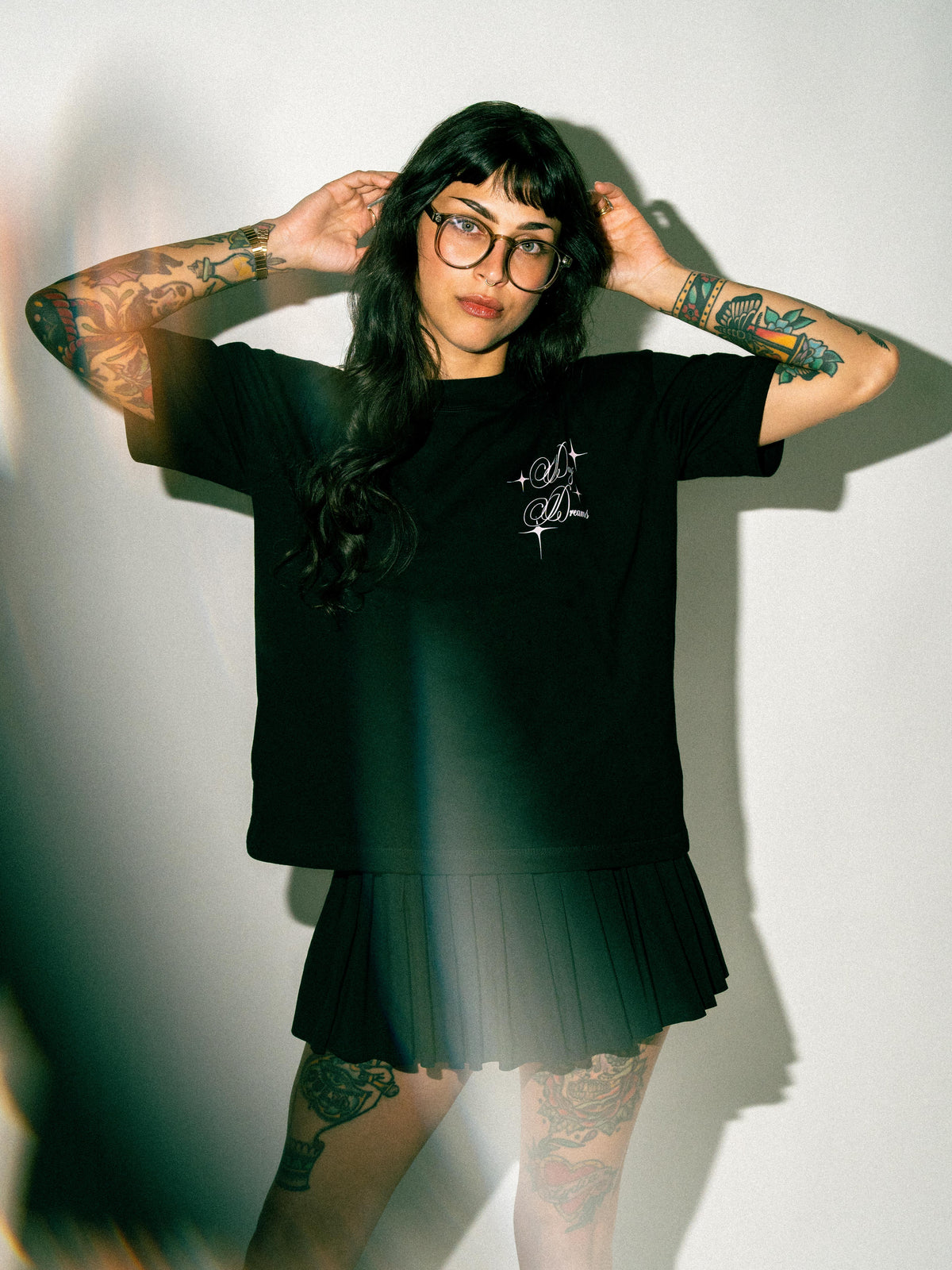 A woman with tattoos and glasses wearing a black t-shirt with a white graphic and a matching black pleated skirt, posing against a white wall with dramatic shadows.