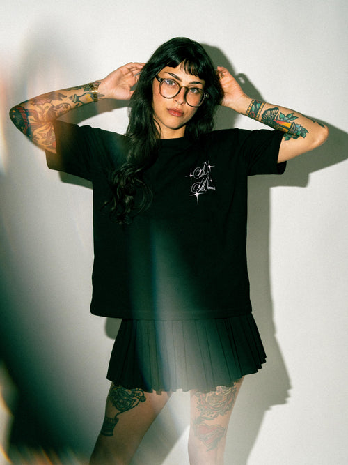 A woman with tattoos and glasses wearing a black t-shirt with a white graphic and a matching black pleated skirt, posing against a white wall with dramatic shadows.