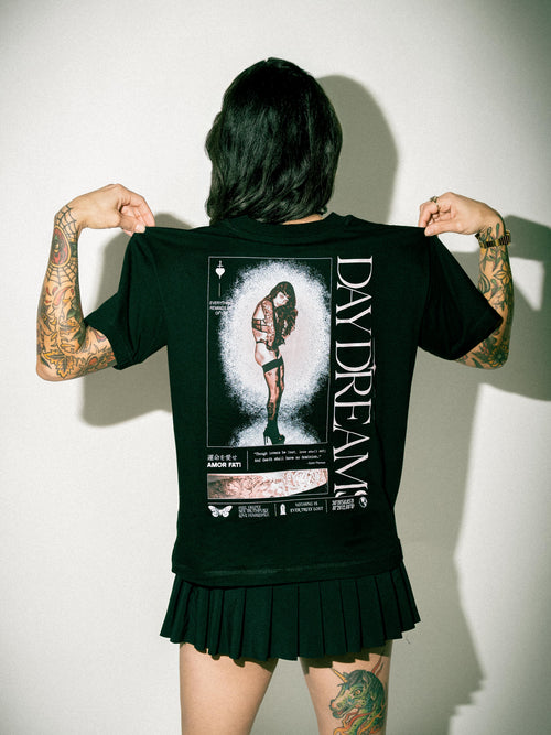 A person shown from behind wearing a black short-sleeve graphic T-shirt and a black pleated skirt, holding the shirt out to show the large back print. The shirt features a white and gray illustrated figure with vertical text reading 'DAYDREAM'.