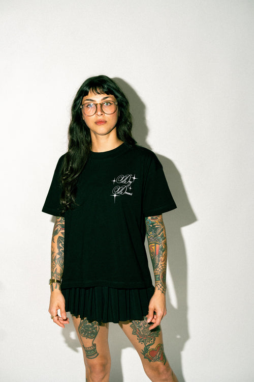 A tattooed model with dark hair and glasses wearing a black oversized t-shirt dress with a pleated skirt hem and a small white graphic on the left chest, standing against a plain white wall.