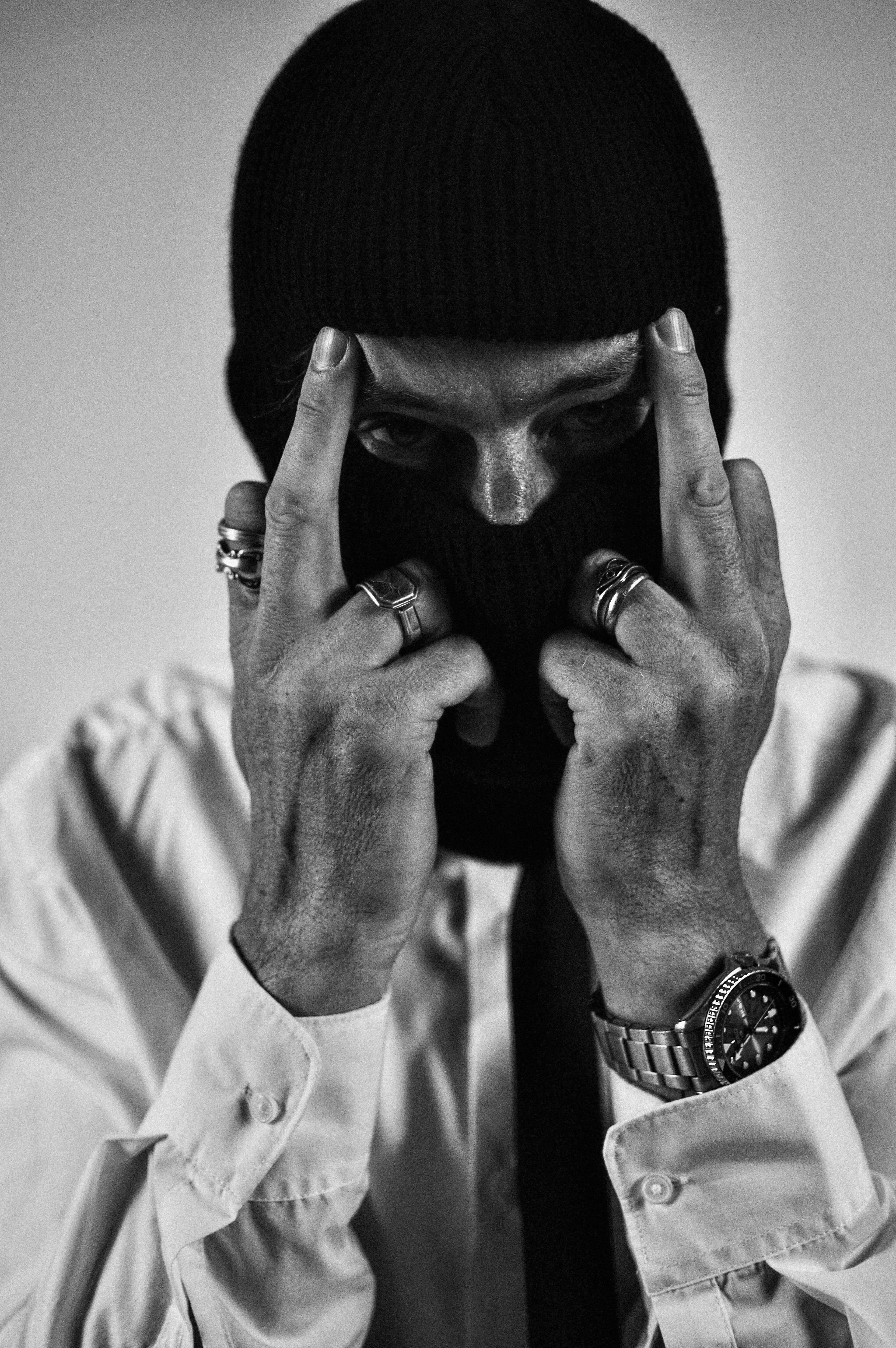 A black and white portrait of a person wearing a black knit beanie and a light-colored button-up shirt, holding their hands up to block part of their face. They are wearing multiple rings and a wristwatch. 
Photo taken by Mikaylah Brooke of Magic by Mikaylah.