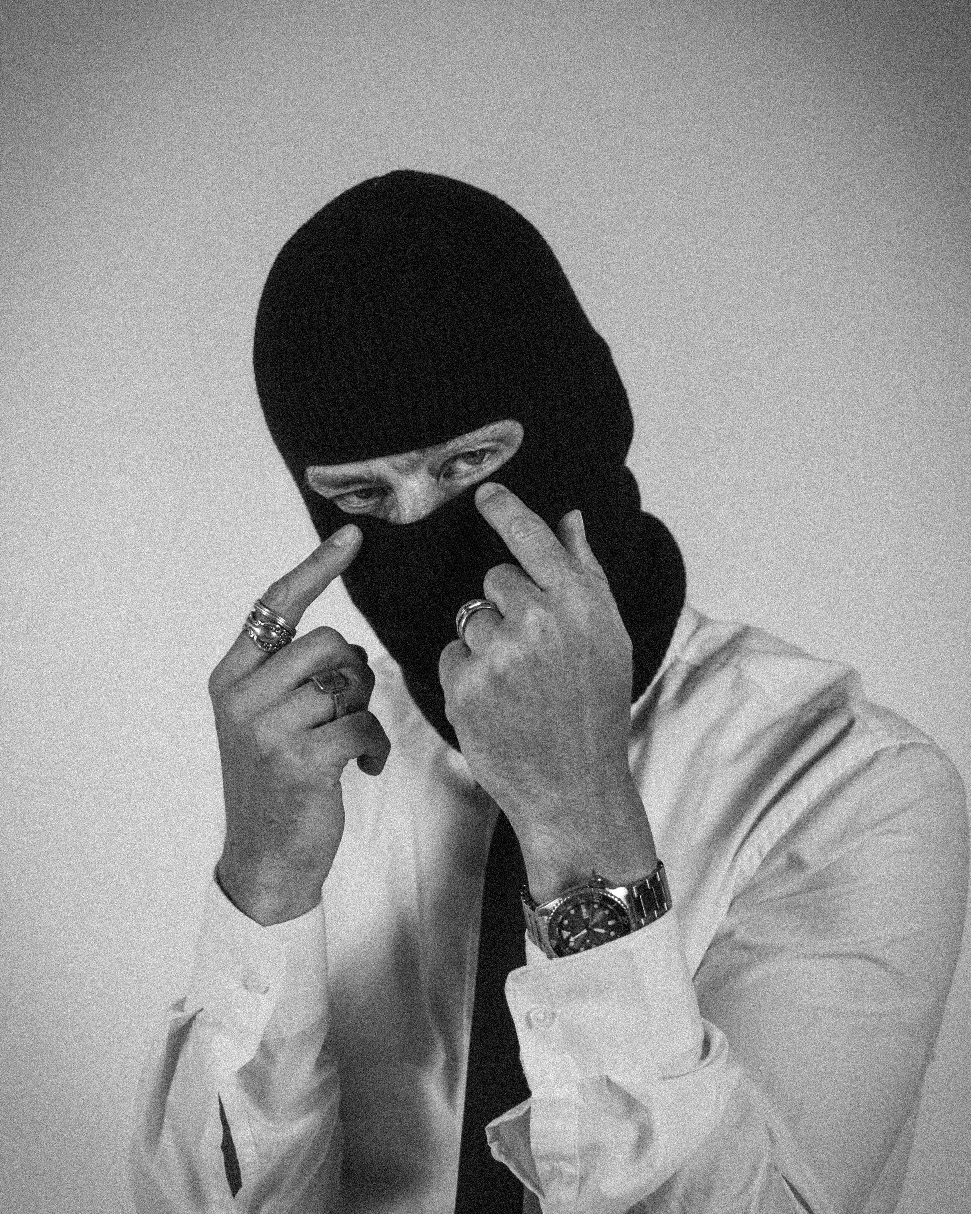 A black knit balaclava mask worn by a person in a white dress shirt and tie, with both hands raised to touch the mask, photographed against a plain light gray background.