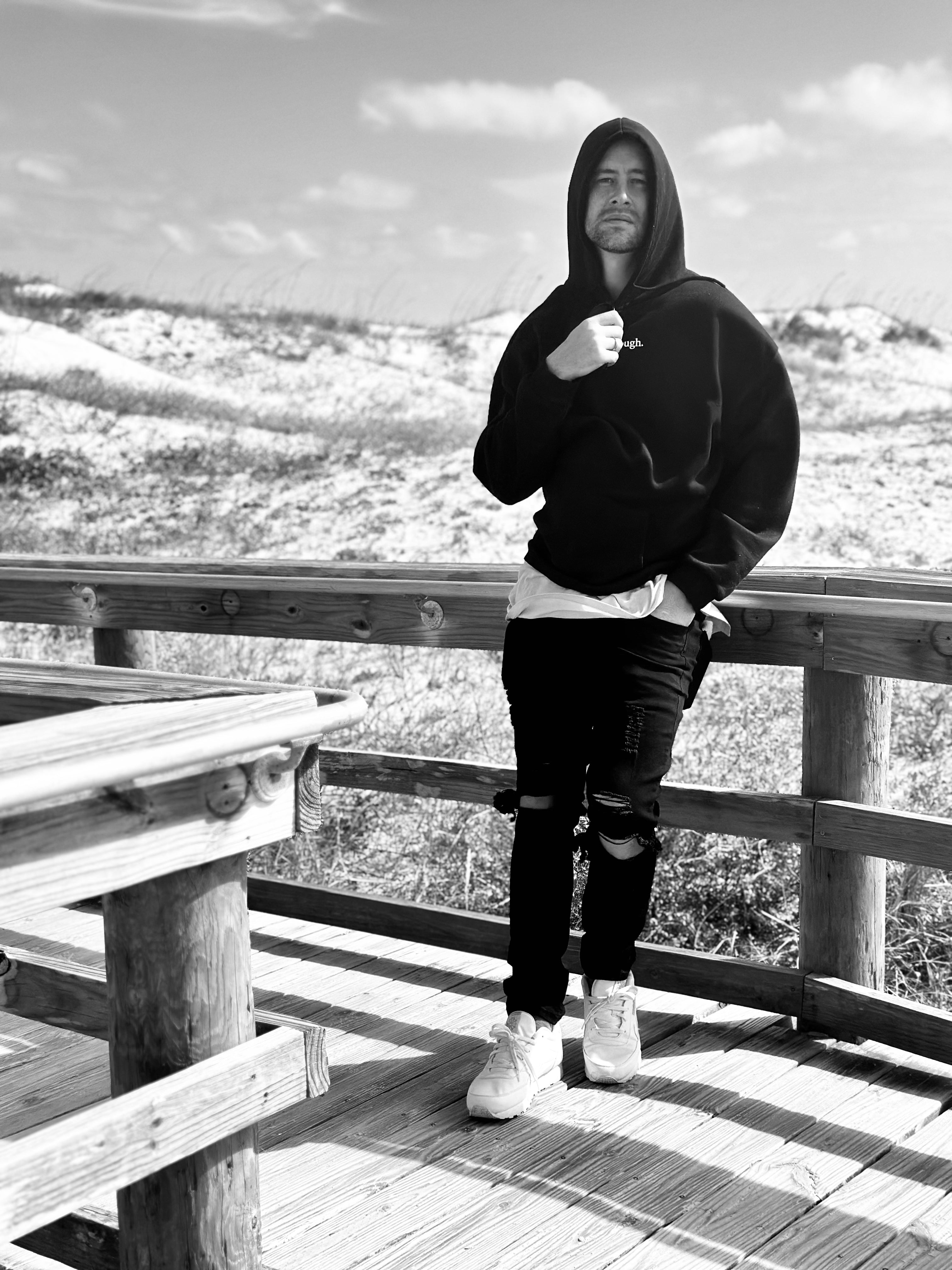A man wearing a black hoodie, ripped black jeans, and white sneakers stands on a wooden boardwalk outdoors in a black and white photograph.