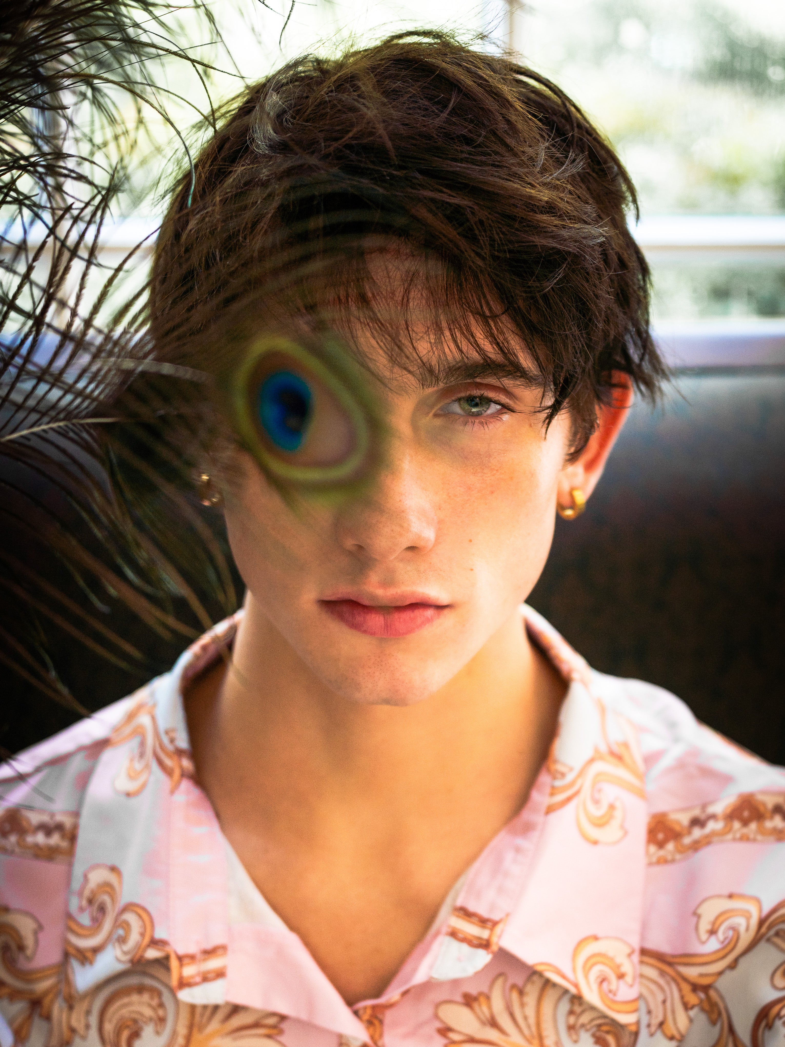 A close-up portrait of a young man with short brown hair wearing a light pink patterned button-up shirt. A peacock feather is draped over his left eye.